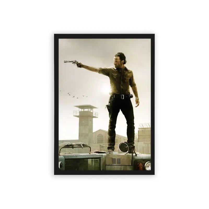 The Walking Dead 'Survivor's Stand' Framed Poster Black Hard Fiber A rugged adventurer stands atop an overturned vehicle, confidently aiming a gun off to the distance, with a watchtower and birds in the dusky sky behind him.