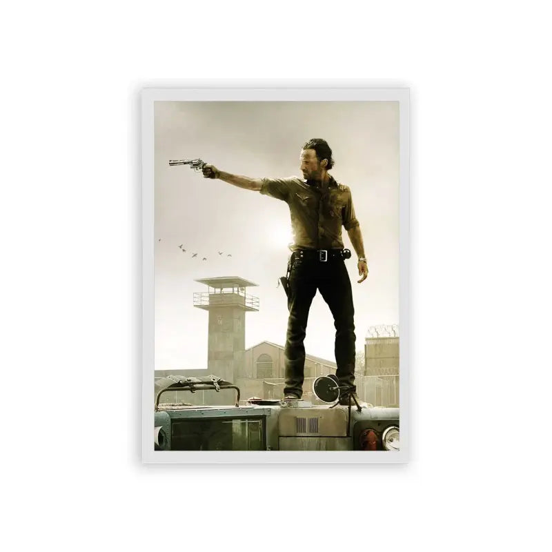 The Walking Dead 'Survivor's Stand' Framed Poster White Hard Fiber A rugged adventurer stands atop an overturned vehicle, confidently aiming a gun off to the distance, with a watchtower and birds in the dusky sky behind him.