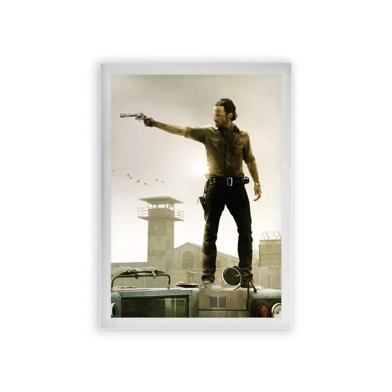 The Walking Dead 'Survivor's Stand' Framed Poster White Premium Wood A rugged adventurer stands atop an overturned vehicle, confidently aiming a gun off to the distance, with a watchtower and birds in the dusky sky behind him.