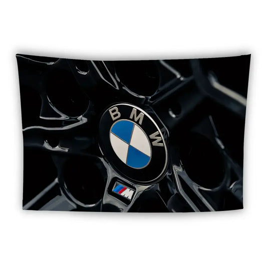 Close-up shot of a BMW logo on a black car grill, featuring the iconic blue and white circular emblem. The M-series badge, representing BMW's high-performance line, is also visible in the lower part of the image. The background is dark, highlighting the logos.