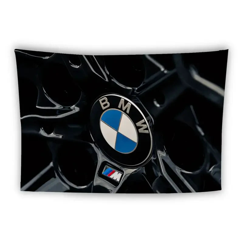 Close-up shot of a BMW logo on a black car grill, featuring the iconic blue and white circular emblem. The M-series badge, representing BMW's high-performance line, is also visible in the lower part of the image. The background is dark, highlighting the logos.