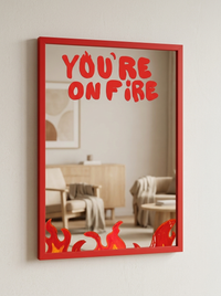 Angled view of red-framed You're On Fire wall mirror with flame illustration in a neutral living room